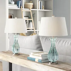 Two elegant table lamps with clear glass bases and white shades are placed on a console table behind a sofa.