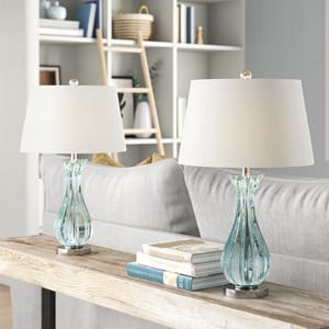 Two elegant table lamps with clear glass bases and white shades are placed on a console table behind a sofa.