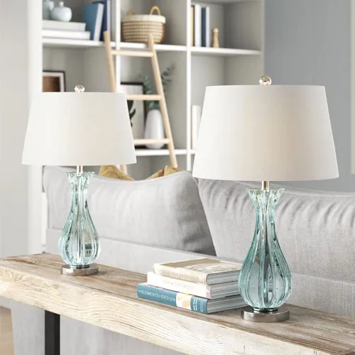 Two elegant table lamps with clear glass bases and white shades are placed on a console table behind a sofa.