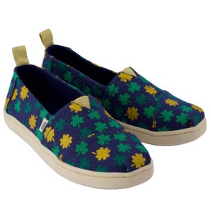 Navy blue slip-on shoes with green and yellow clover patterns, featuring a white sole and gold accents on the tongue and heel loops.