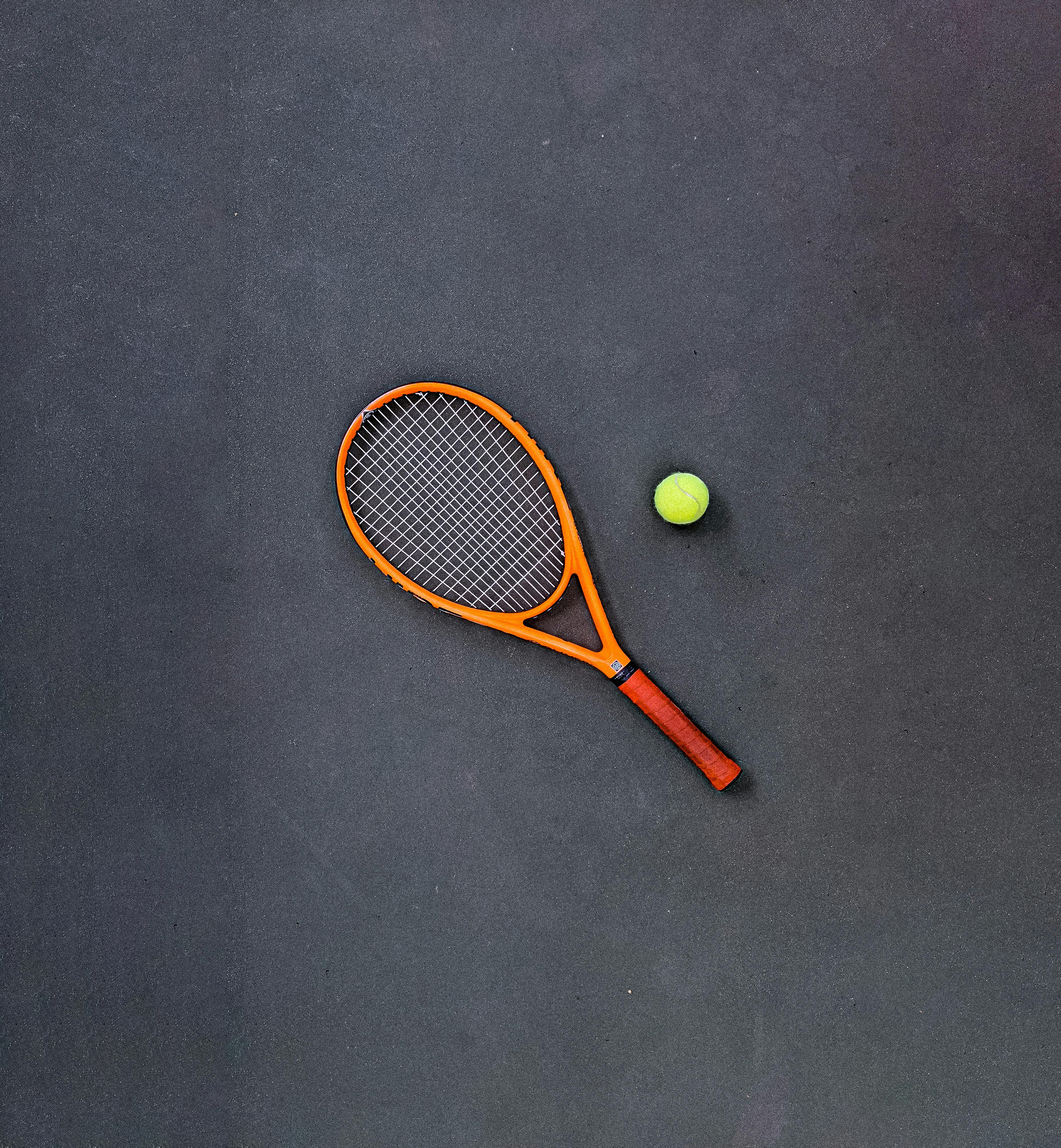 Orange and black tennis racket with a yellow tennis ball on a dark surface.