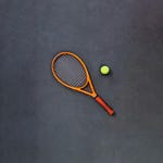 Orange and black tennis racket with a yellow tennis ball on a dark surface.