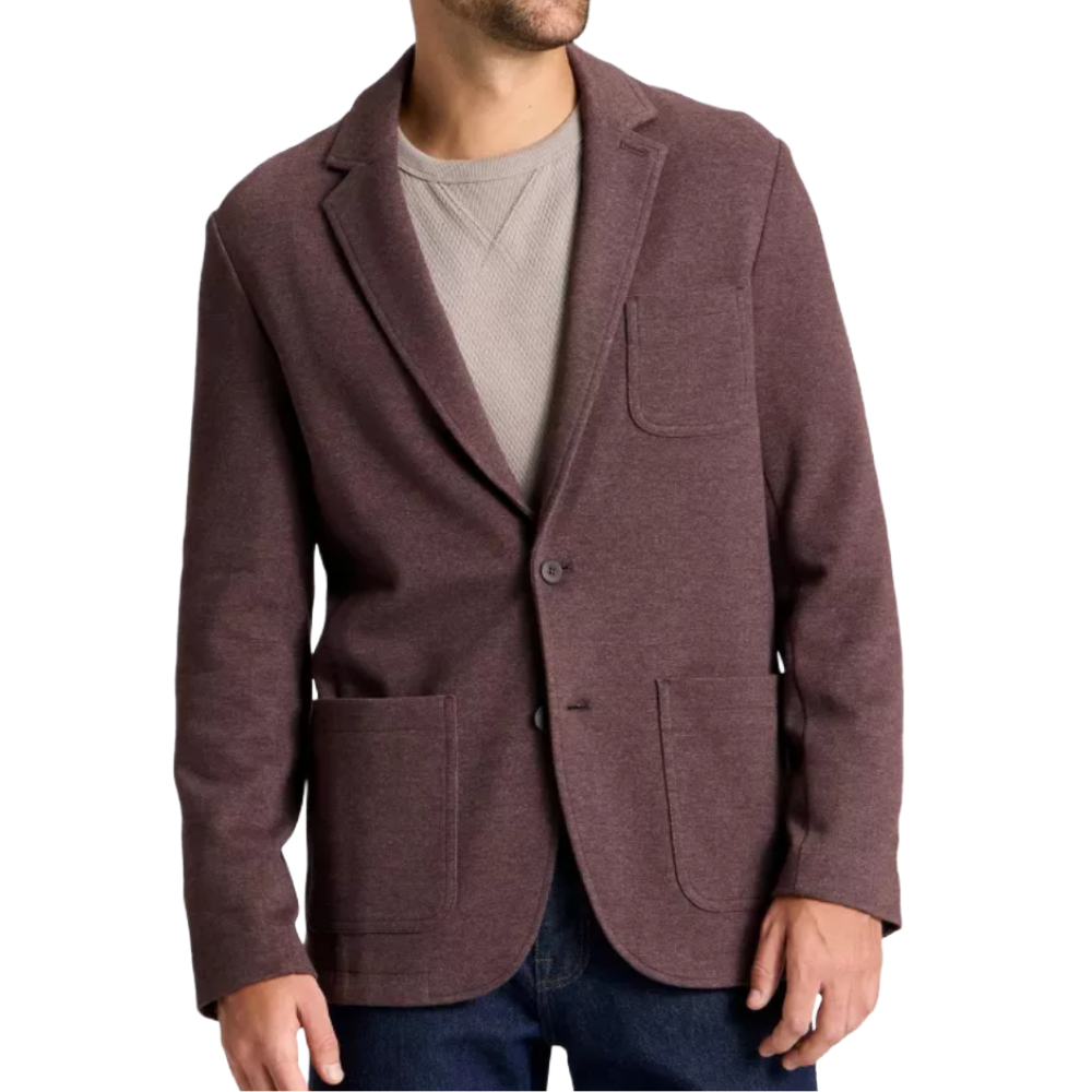 A man is wearing a maroon two-button blazer with patch pockets, layered over a grey textured sweater, paired with dark jeans.