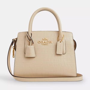 A beige, crocodile-embossed leather carryall with dual top handles, a detachable strap, and gold-tone Coach branding and hardware accents.