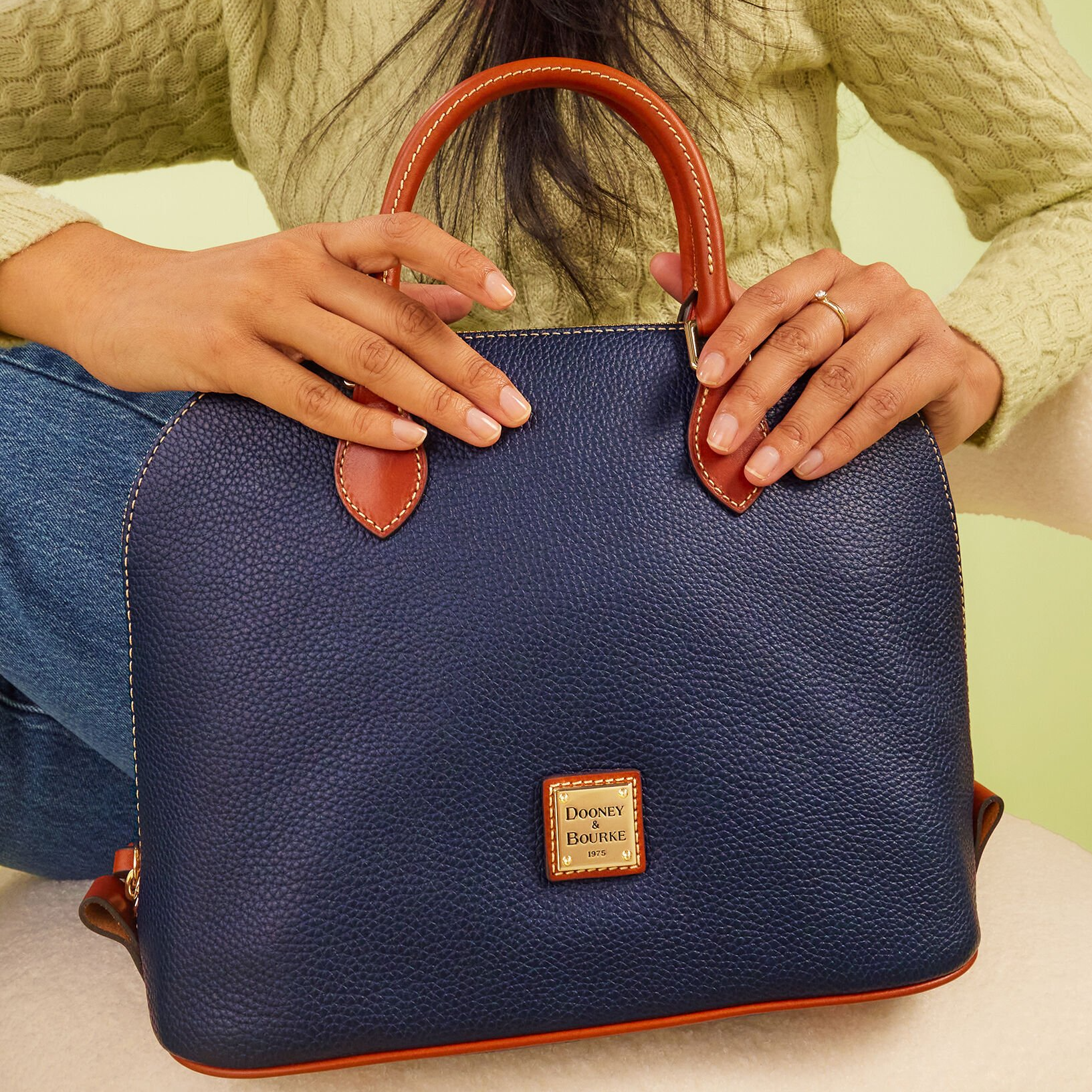 A navy blue Dooney & Bourke pebble leather domed satchel with brown leather handles and accents, featuring a gold-tone logo plate on the front.