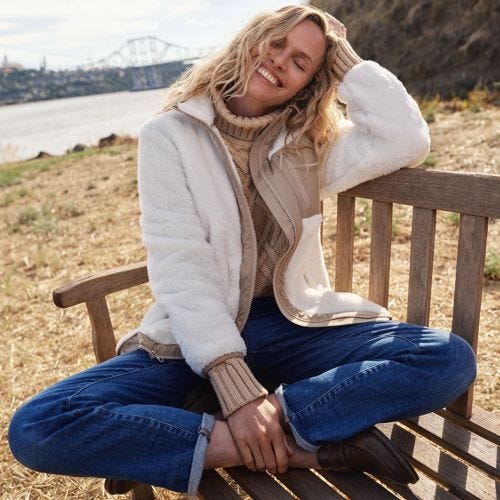 A woman is seated wearing a white Sherpa fleece jacket with a zipper front, side pockets, and a standing collar. She's also wearing a beige sweater and blue jeans.