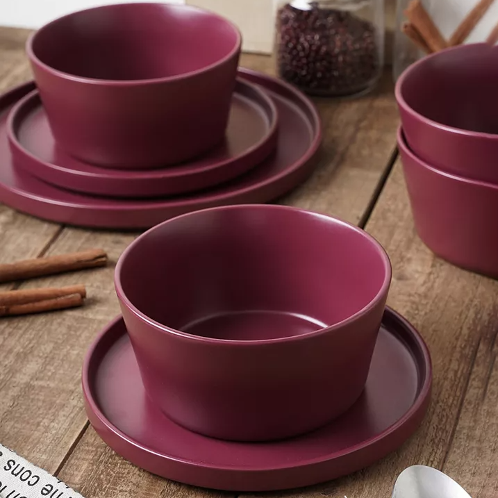 A 24-piece stoneware dinnerware set in burgundy, including plates, bowls, and saucers, displayed on a wooden table.