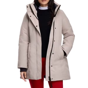 A woman is wearing a light beige winter coat with a hood, over a black sweater and white shirt. She pairs it with red pants.