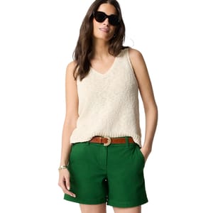 A woman wearing a sleeveless beige knit top, green shorts with a brown belt, and dark sunglasses.
