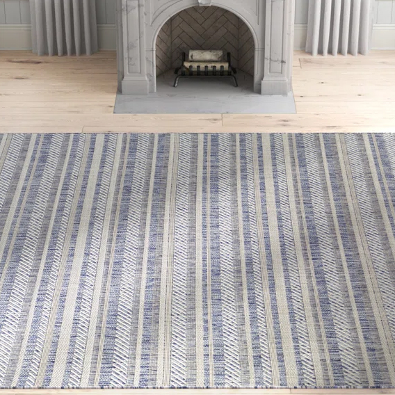 A striped rug featuring blue and beige tones is placed on a light wooden floor in front of a decorative gray fireplace.