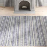 A striped rug featuring blue and beige tones is placed on a light wooden floor in front of a decorative gray fireplace.