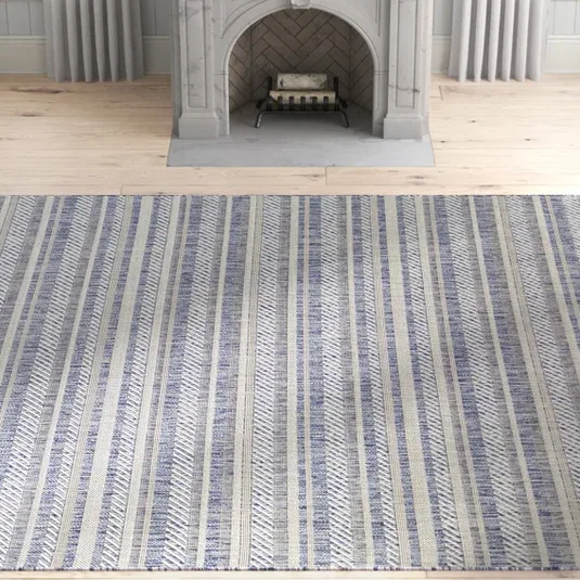 A striped rug featuring blue and beige tones is placed on a light wooden floor in front of a decorative gray fireplace.