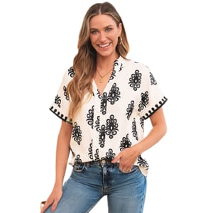A short-sleeve V-neck blouse features an ivory backdrop with a symmetrical black ornate pattern and black trim along the sleeves. The blouse is paired with blue jeans.