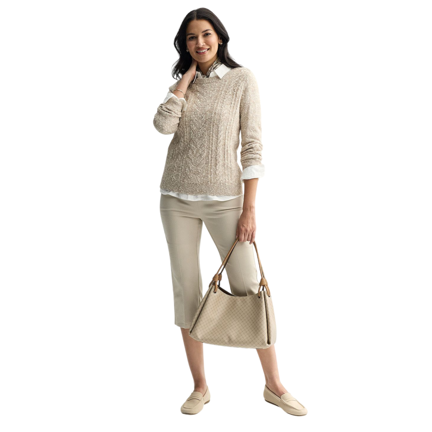 A woman is wearing a beige cable-knit sweater over a white shirt, cropped beige pants, and beige loafers, carrying a beige textured handbag. She has long dark hair and is smiling.