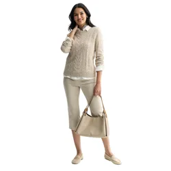 A woman is wearing a beige cable-knit sweater over a white shirt, cropped beige pants, and beige loafers, carrying a beige textured handbag. She has long dark hair and is smiling.