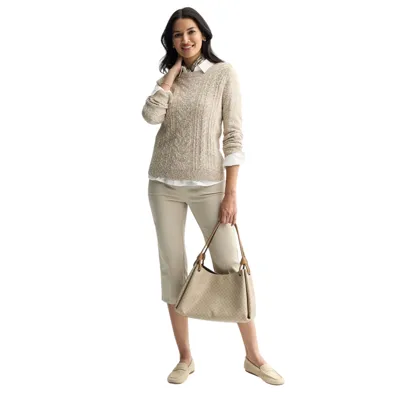 A woman is wearing a beige cable-knit sweater over a white shirt, cropped beige pants, and beige loafers, carrying a beige textured handbag. She has long dark hair and is smiling.