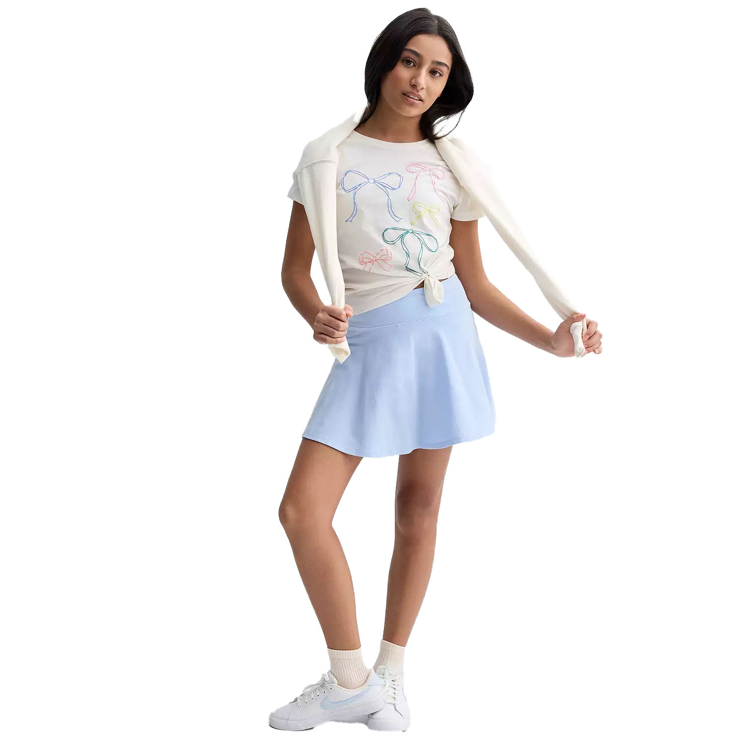 A woman is wearing a white t-shirt with colorful bow designs, a light blue skirt, and white sneakers, with a white sweater draped over her shoulders.