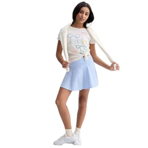 A woman is wearing a white t-shirt with colorful bow designs, a light blue skirt, and white sneakers, with a white sweater draped over her shoulders.
