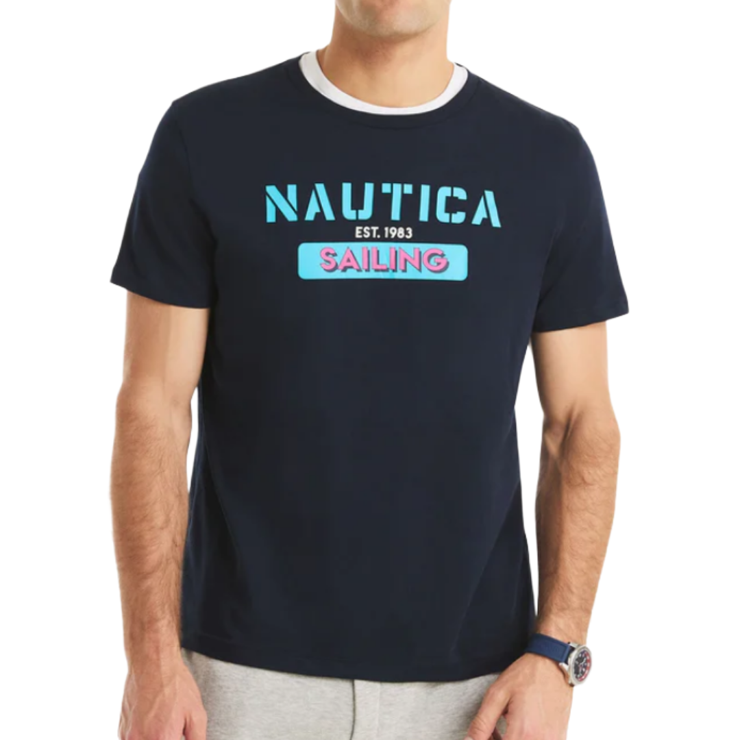 A navy blue T-shirt with \“NAUTICA EST. 1983\“ and \“SAILING\“ printed on the front in blue and pink. The model is wearing a watch and gray pants.