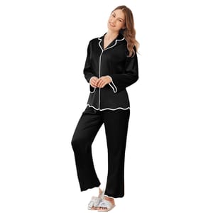 Women's satin pajamas in black with white piping, featuring a long-sleeve button-up top and matching pants. The outfit is completed with open-toe sandals adorned with bows.