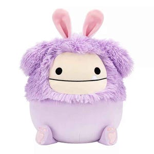A plush Squishmallows character with a round shape, lavender body, fuzzy purple fur, floppy ears, and simple black eyes. It has soft features and resembles a playful, cute creature.