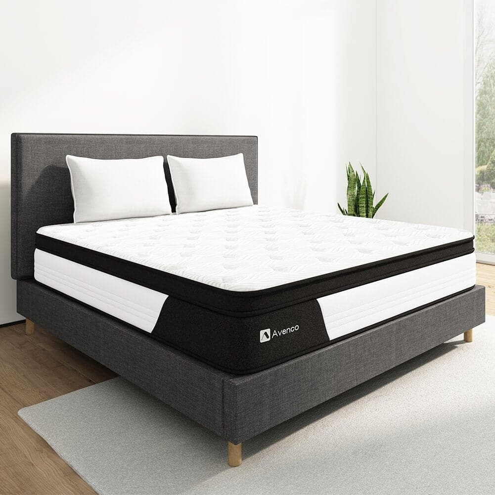 A modern bed with a white and black Avenco mattress, two white pillows, a dark gray upholstered frame, and a small plant beside.