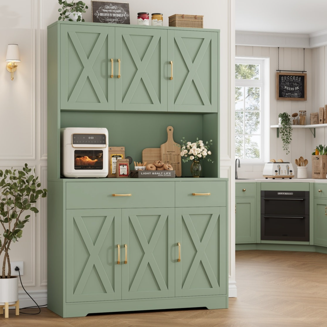 A kitchen features sage green cabinets with X-patterned doors, brass handles, and a countertop displaying an air fryer, cutting boards, and decor items. A corner section includes a black oven, white toaster, and shelves with jars and plants.