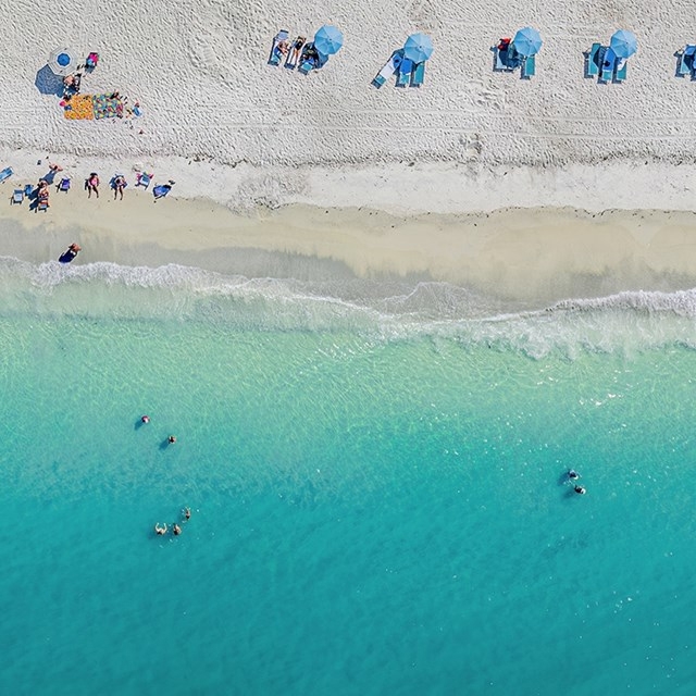 Aerial view of a beach scene featuring multiple blue umbrellas on the sand, with people relaxing under them and swimming in the turquoise water.