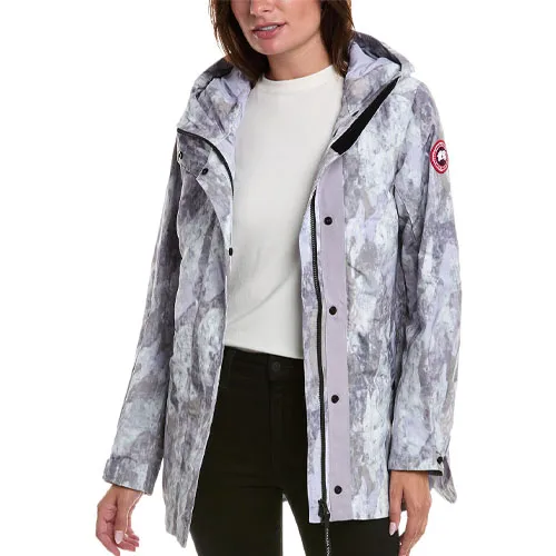 A woman models a Canada Goose Mindin Jacket featuring a light-colored, abstract pattern with a hood, front snap closure, and logo patch on the left arm. She wears a white top and black pants.
