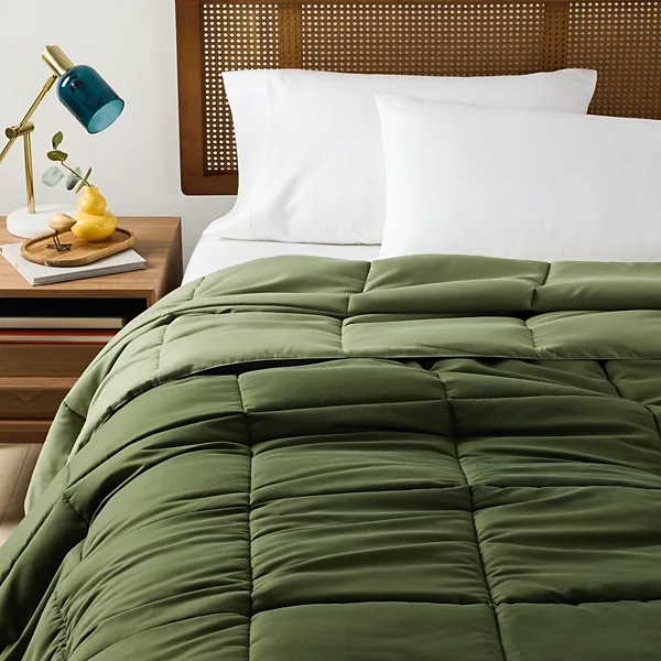 Green microfiber down-alternative comforter with square quilted pattern, displayed on a bed with white pillows and a wicker headboard. A wooden nightstand with a blue lamp and decorative items is nearby.