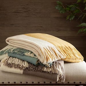 A stack of three fringed throw blankets in varying shades of beige, yellow, and blue rests on a beige ottoman.