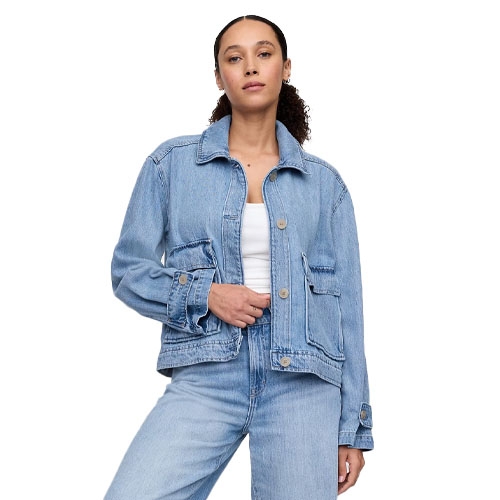 A woman is wearing a light blue denim jacket with a relaxed fit, featuring large pockets and button closures. The jacket is paired with matching denim jeans, and underneath is a simple white top.