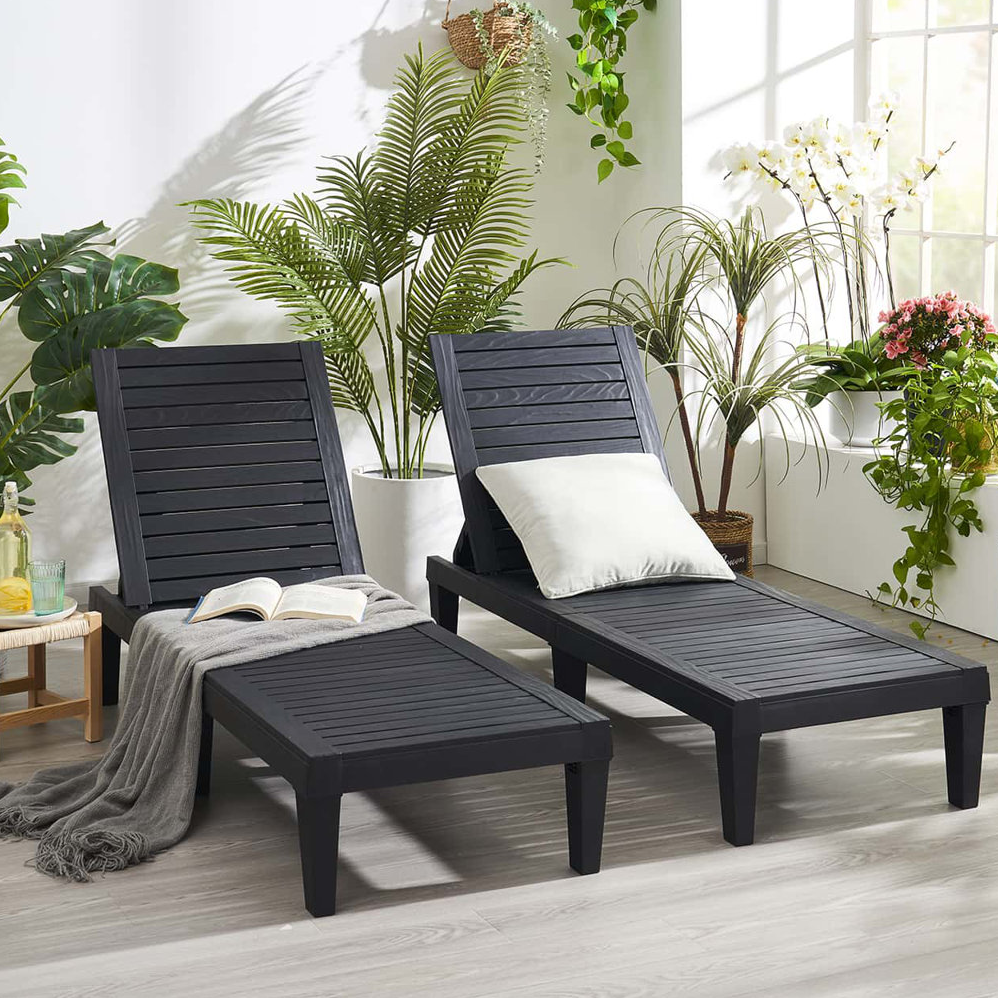 Two black outdoor reclining lounge chairs with a cushion and a blanket. Surrounded by various potted green and flowering plants, there is also an open book and a refreshing drink placed on a side table.