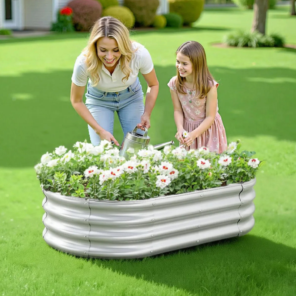 A woman and a girl are watering white flowers in a raised metal garden bed on a green lawn.