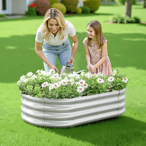 A woman and a girl are watering white flowers in a raised metal garden bed on a green lawn.