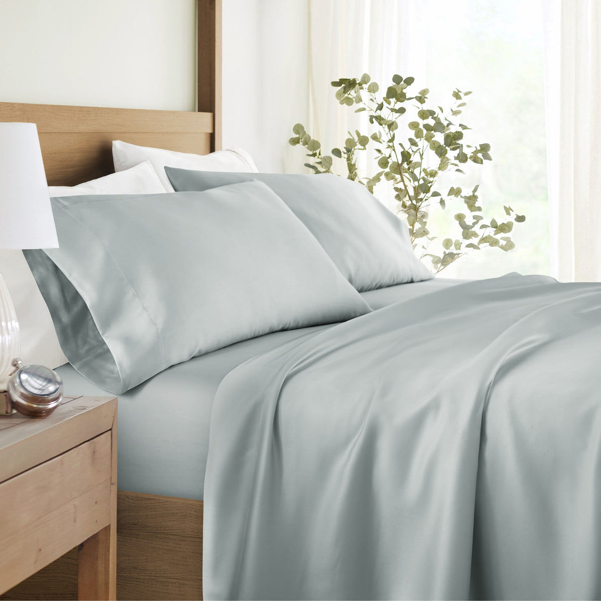 A bed with light gray satin sheets and pillowcases, placed against a wooden headboard. A plant is visible beside the bed.