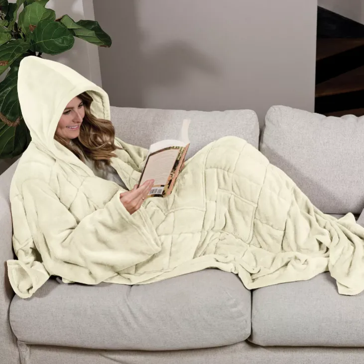 A person relaxes on a couch wearing a hooded blanket while reading a book. The blanket has a quilted design and covers the entire body.