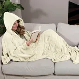 A person relaxes on a couch wearing a hooded blanket while reading a book. The blanket has a quilted design and covers the entire body.