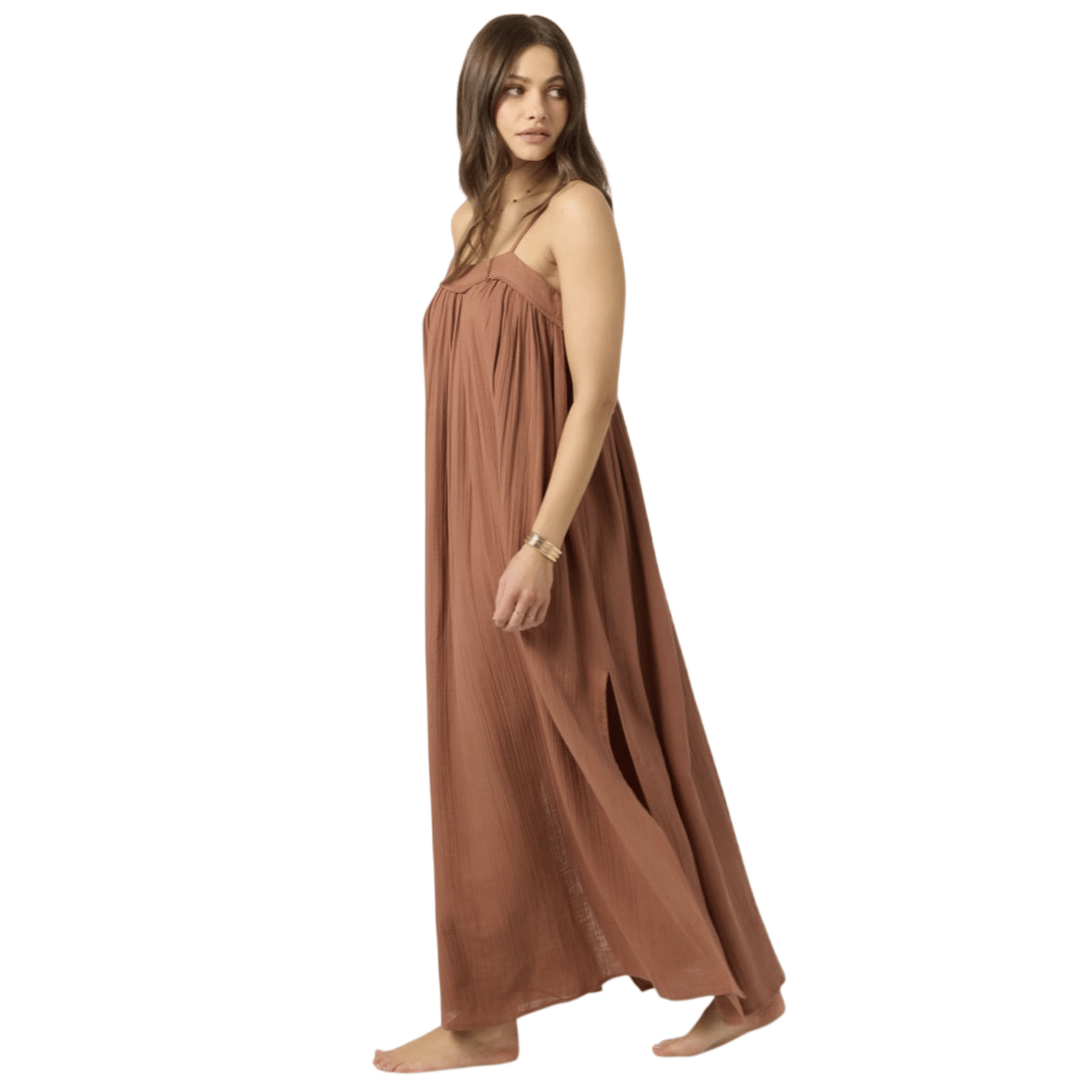 A woman is wearing a flowy, brown maxi dress with thin straps and a side slit, accessorized with a bracelet.