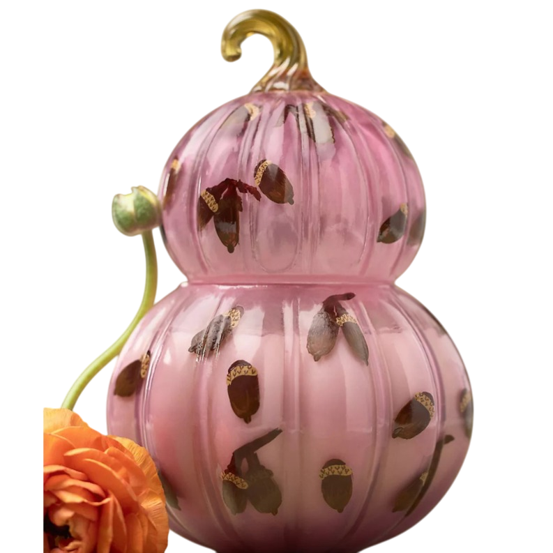 A pink, gourd-shaped glass candle decorated with brown and gold acorn designs next to a vibrant orange flower.