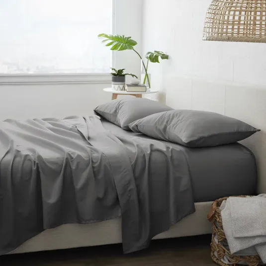 A bedroom scene featuring gray bed sheets and pillows, a small side table with potted plants, and a wicker basket with linens, creating a cozy and minimalist aesthetic.