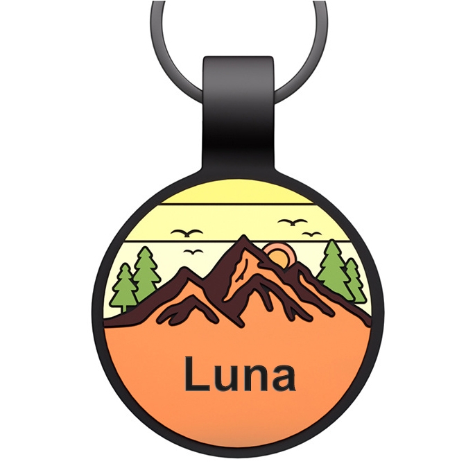 Round pet ID tag with a nature scene, featuring mountains, trees, and birds against a yellow-orange sunrise background, and the name \“Luna\“ displayed at the bottom. The tag has a black border and loop for attachment.