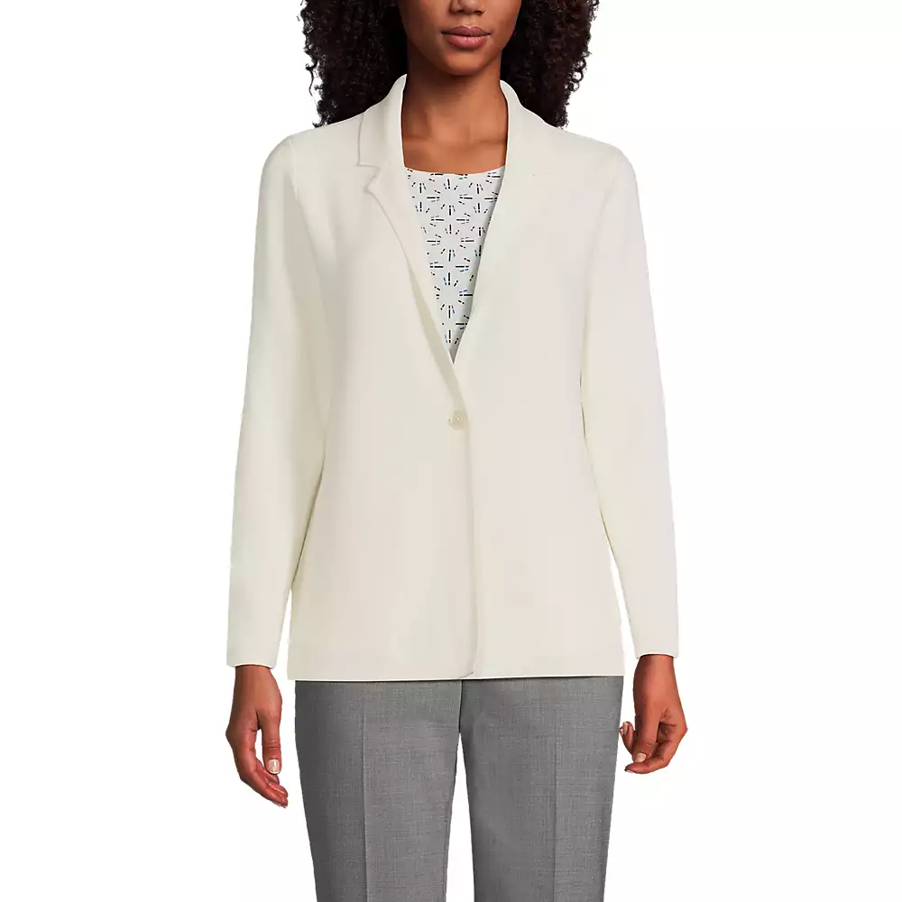 A woman wears a white blazer over a patterned top, paired with gray trousers.