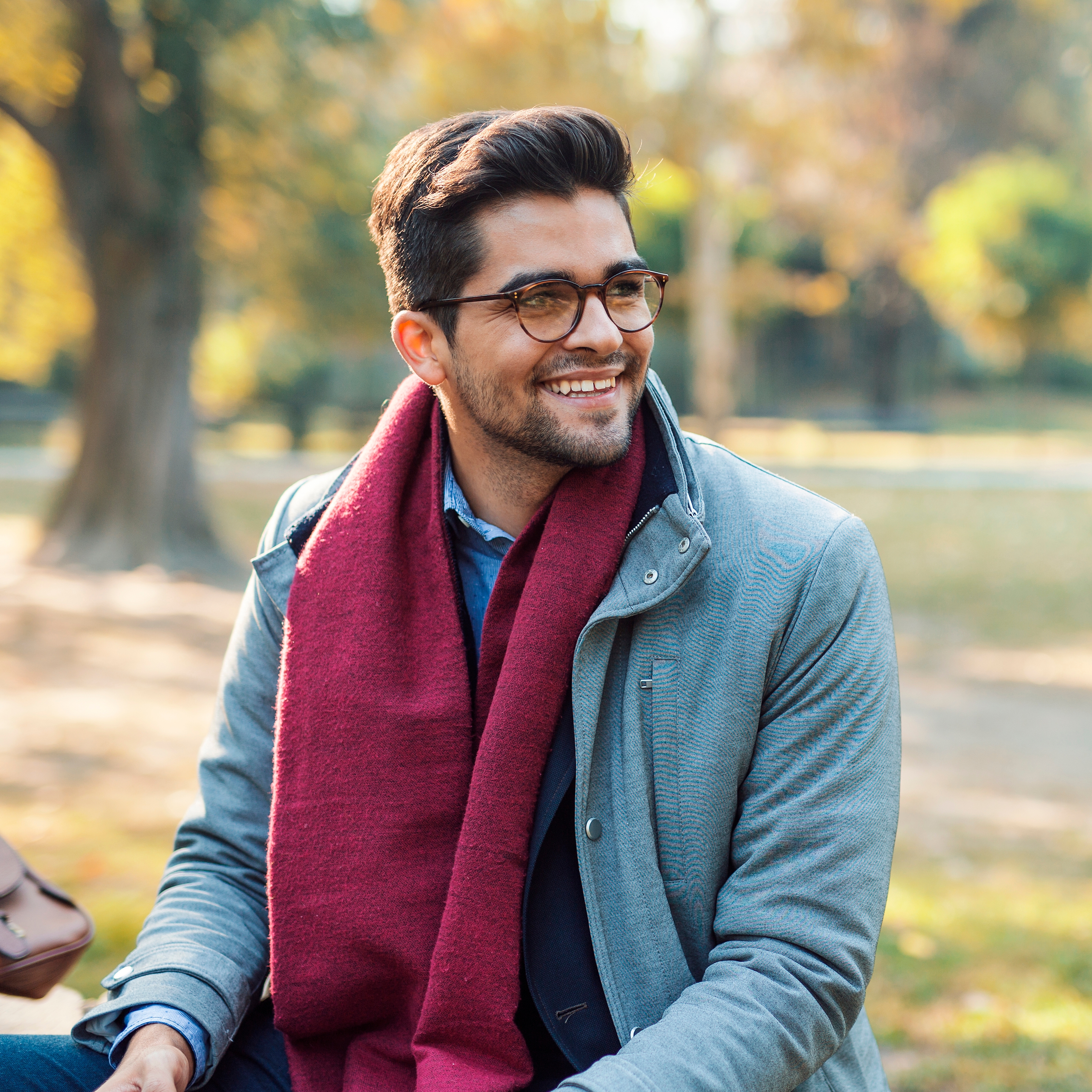 A smiling man wearing glasses, a grey coat, dark scarf, and a blue shirt.