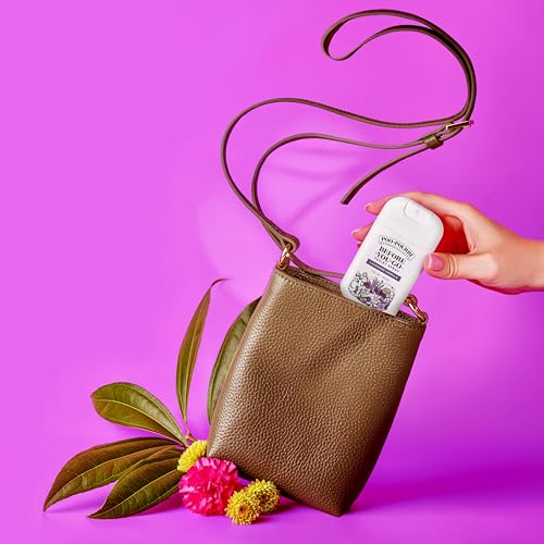 A hand places a lavender and sage scented deodorant from Schmidt's into a brown leather crossbody bag surrounded by leaves and colorful flowers on a vibrant purple background.