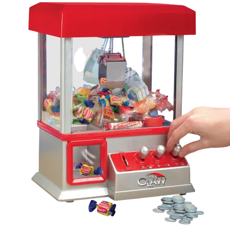 A miniature claw machine filled with assorted candies, featuring a red and silver design and joystick controls. It is accompanied by a collection of tokens for operation.