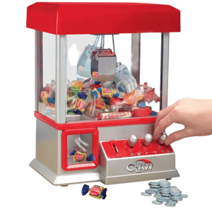 A miniature claw machine filled with assorted candies, featuring a red and silver design and joystick controls. It is accompanied by a collection of tokens for operation.