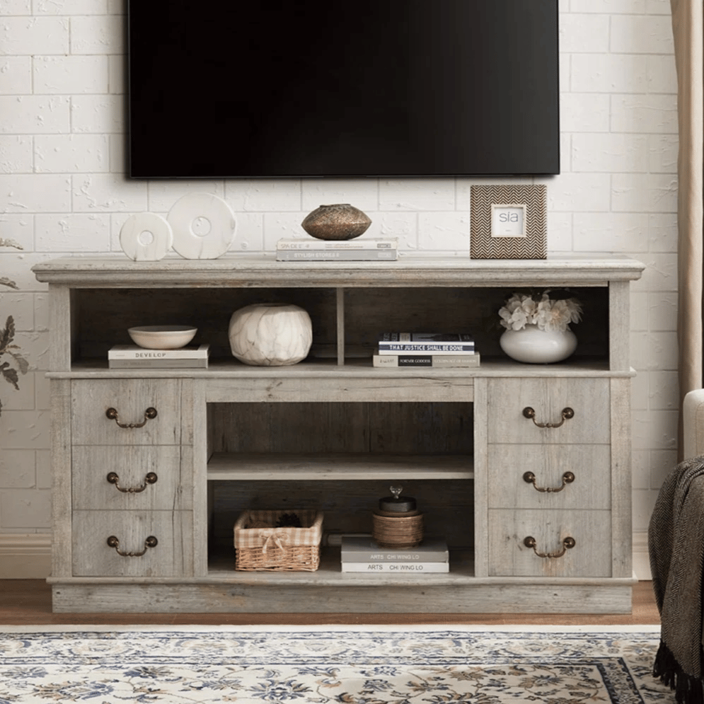 A rustic wooden TV stand with six drawers and multiple open shelves holds decor items like books, bowls, and vases. A large flat-screen TV is mounted above, and a floral rug lies on the floor.