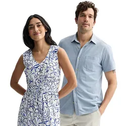 A woman in a sleeveless, floral-patterned, V-neck dress stands beside a man wearing a short-sleeve, light blue button-up shirt paired with light-colored pants.
