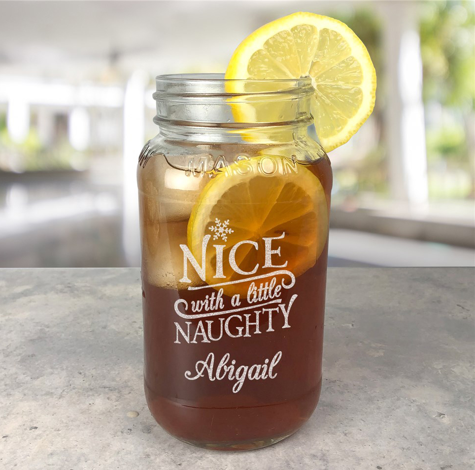 A mason jar filled with iced tea and lemon slices features an engraved message: \“Nice with a little Naughty, Abigail,\“ with a lemon wedge on the rim.